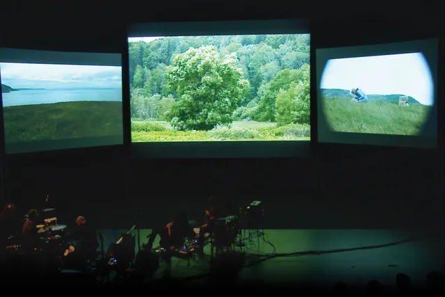 Three screens showing greenery and rolling hills behind a small pit orchestra on a dark stage.