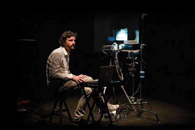 A white man with dark hair sitting cross legged next to a slide projector and lap top. 