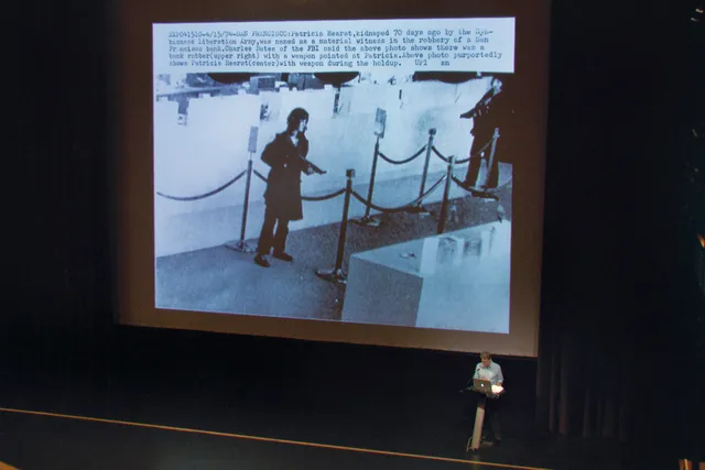 Thomas Keenan on stage in front of a large projection of an image of Patricia Hearst robbing a bank 