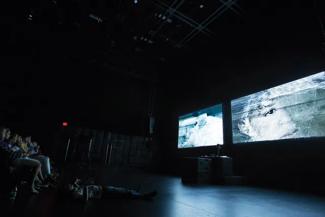 Two screens projecting scratched images of human eyes to a seated audience in a black theater.