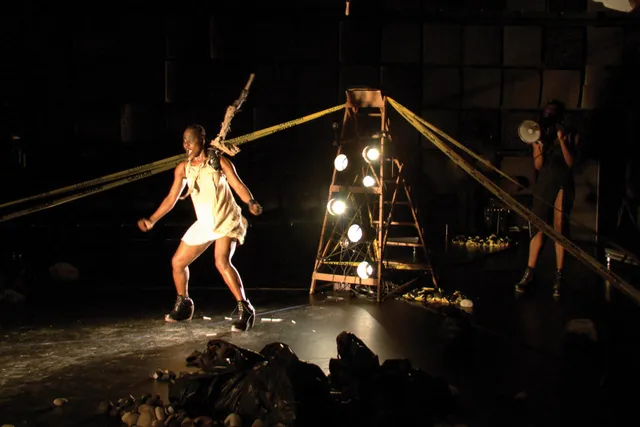 A Zimbabwean woman on a harness dancing in black box studio amongst piles of trash bags, caution tape, and a ladder with stage lights placed on the steps.