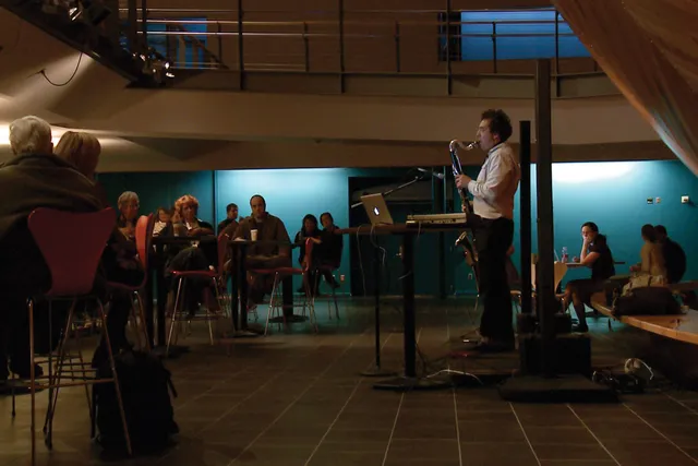 David Rothenberg playing saxophone to a small audience seated in a cafe inside of EMPAC. 