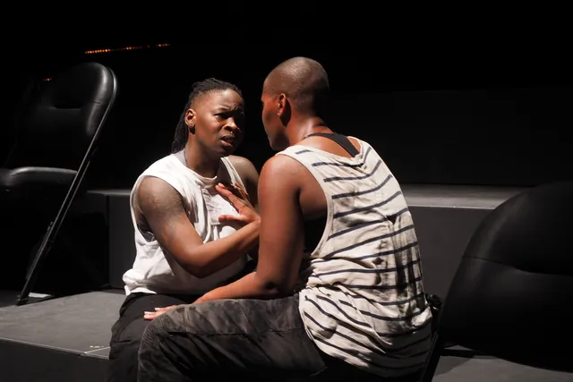 Two Black woman sitting facing each other on the steps of a black box theater.