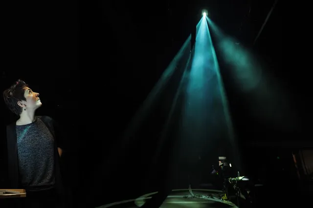 Nina Young looking up dramatically at a blue beam of light o a dark stage.