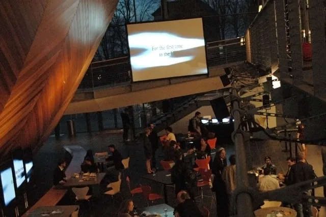 A reception full of people milling about under the hull of the EMPAC concert hall. A screen projecting the words "For the first time in the.." is suspended above the crowd. 