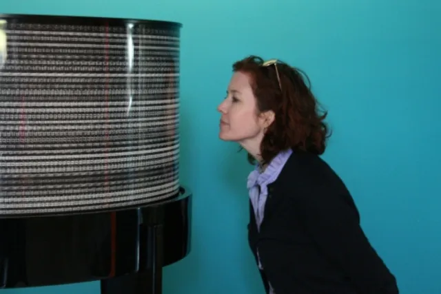 Kathleen Forde leaning towards a black and grey striped cylinder against a teal wall. 