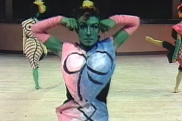 A woman painted green wearing a half pink, half blue costume with a Picasso inspired nude female form painted on her outfit. Two other female performers are in the background similarity dressed.  
