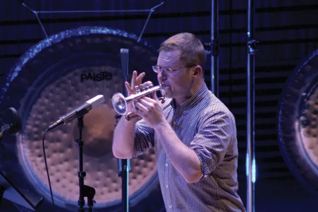 A white male playing a soprano trumpet into a microphone in front of a gong.