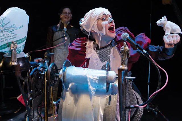 A woman draped in white slime standing in front of a spindle of plastic sheeting speaking theatrically to a white bobble head dog.