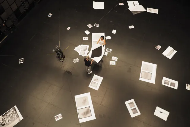 A man and a woman holding a large white piece of paper with a black diamond amongst other papers of various sizes strewn about the floor of a black box studio 