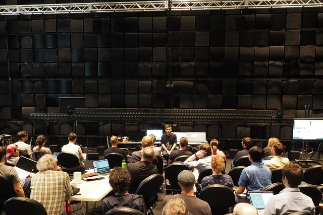 A white male wearing a black t-shirt and jeans sitting on a stool in a room with various computer set ups addressing a packed crowd, attentively looking on in a black box studio.