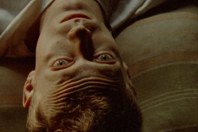 A white man with blonde hair laying on a stained striped pillow looking up at the camera, pensively.