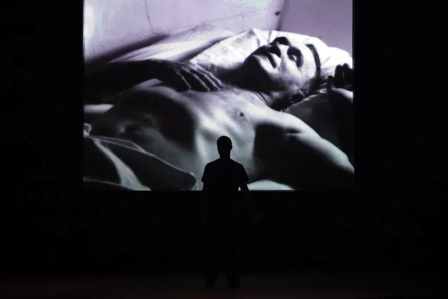 A man silhouetted against a large projection of a shirtless man sleeping. 