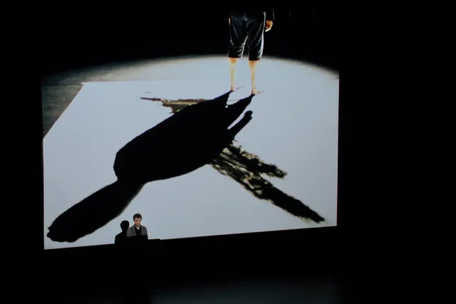 A large screen displaying the shadow of a man wearing shorts and standing in the upper right corner.