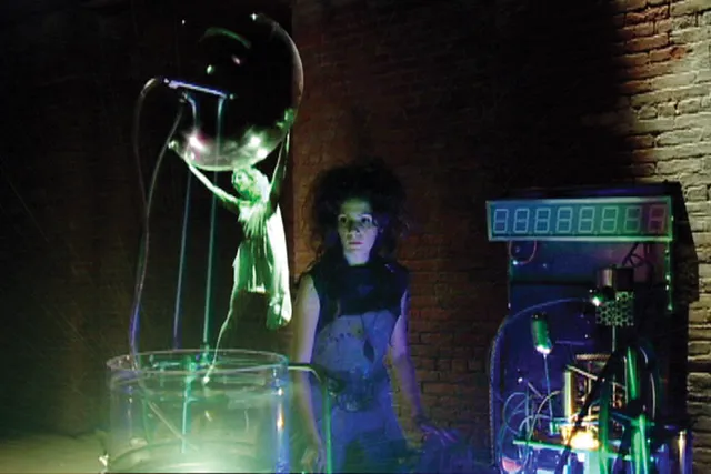 A woman standing amongst various electronic equipment looking at a projection of a small female spectre. 