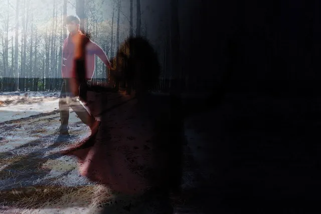 A double exposed image of a shadow of a woman reaching and a man wearing a red jacket walking through a sun dappled forest.
