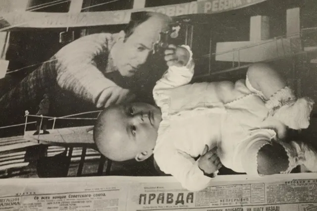 A vintage black and white Russian ad in a newspaper of a balding man reaching across a mechanical item. A baby wearing a sweater and booties is laying on top of the paper.  