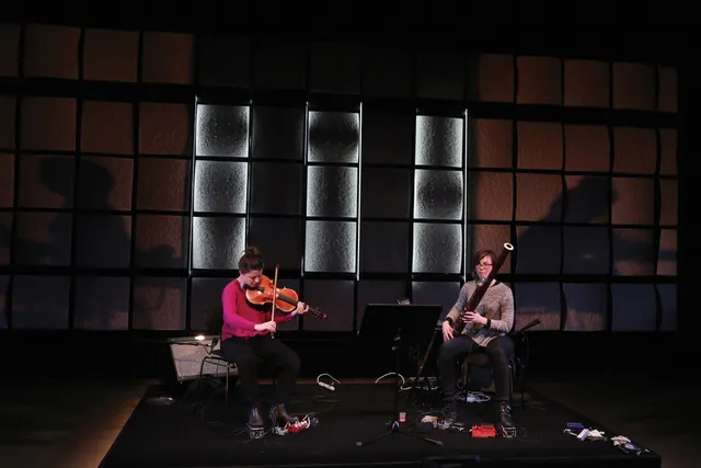 two woman on stage playing instruments in a black box studio. 