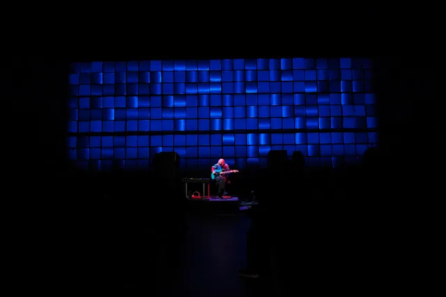 Ellen Arkbro playing an electric guitar on stage while washed in pink light in front of a grid of blue squares.