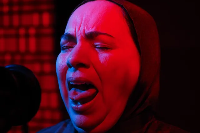 a muslim woman singing into a microphone washed in red light. 