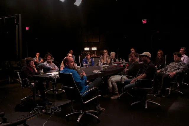 A large group of people gathered in discussion around a conference table in a darkened room.