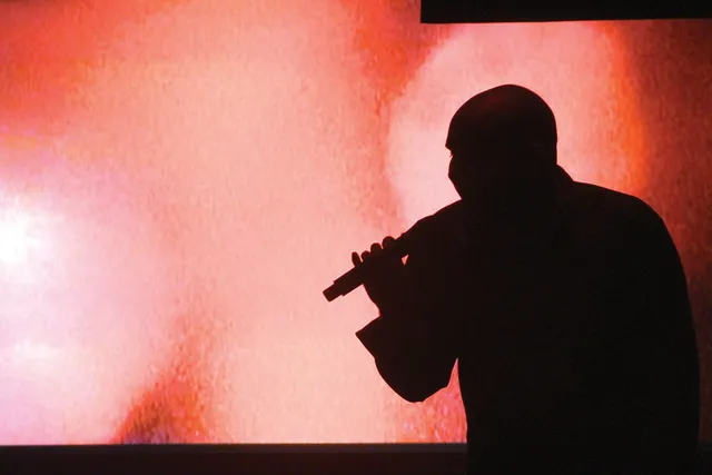A bald person silhouetted against a red watercolored background holding a microphone. 