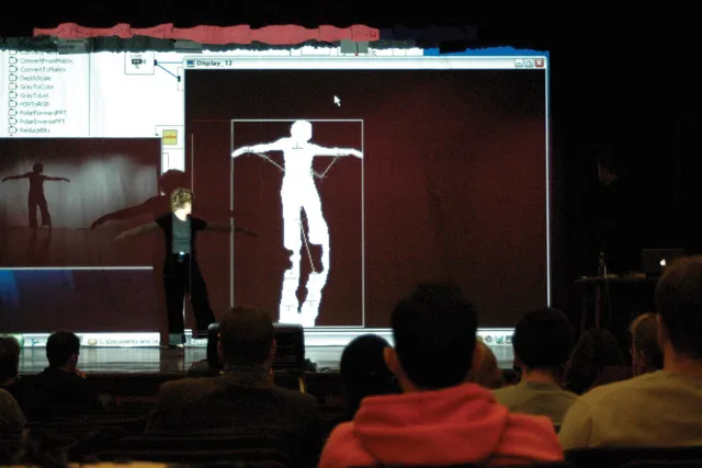A woman standing on stage with arms outstretched as her image is projected on to multiple screens behind her. A full audience looks on. 