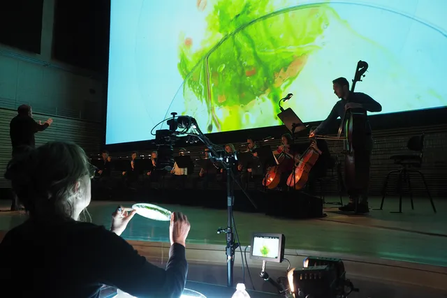 An orchestra on stage in front of a projected image of an lime green blob that is being controlled by a woman in the foreground.