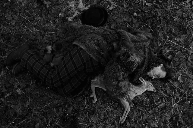 A man wearing a fur coat and tartan pants laying on the ground next to a deceased fawn in a cuddling position.