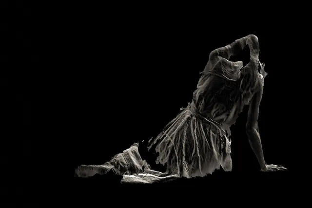 The specter of a woman wearing ragged clothes sitting on the floor with back to the viewer leaning on one arm with the other up over her head.