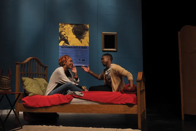 A Black woman and a Black man in a blue room sitting on a bed with red sheets in animated joyous conversation.