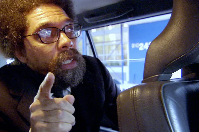 A Black man with an afro and goatee sitting in the back of a car pointing his finger in conversation to the unseen driver. 