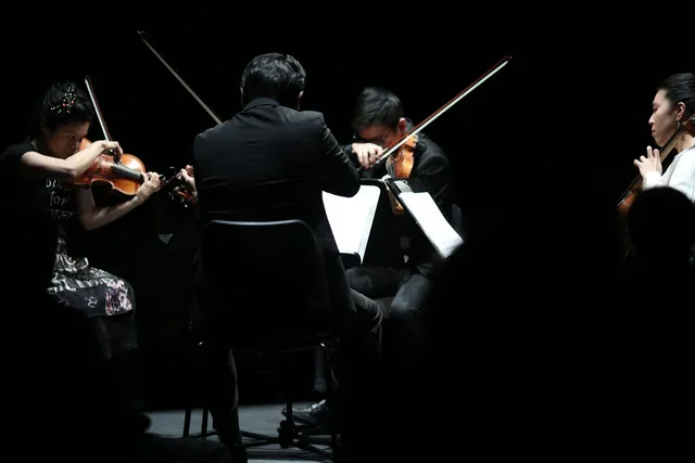 Formosa Quartet actively playing in concert under a spotlight on a dark stage. 