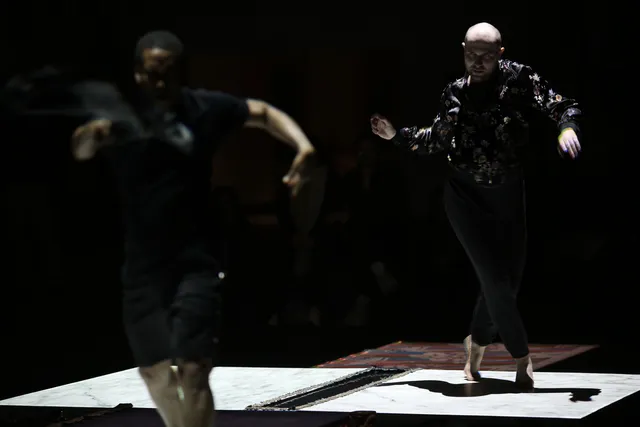 Two male dancers voguing across a stage light by squares of white light on the ground.