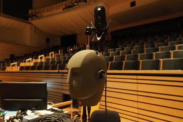 A robotic humanoid head suspended under a microphone in front of theater seating. 