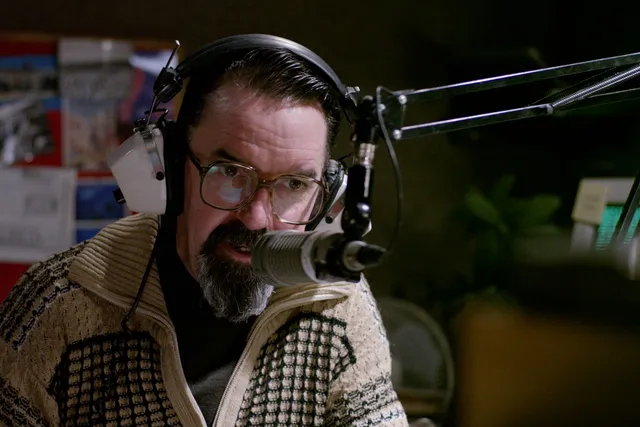 Gerard Byrne speaks into a 70's era radio microphone while wearing white vintage style headphones.