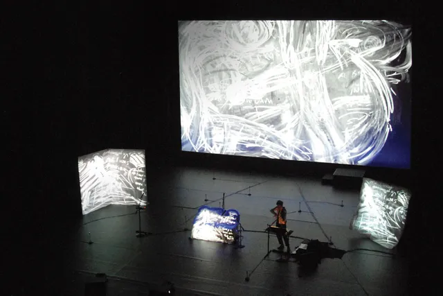 Laurie Anderson in lecture surrounded by screens of various sizes projecting images of chalk swirls. 