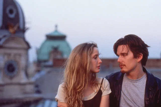 A woman with blonde hair and a man with brown hair looking longingly into each other eyes while sitting on a roof top overlooking a European cityscape.