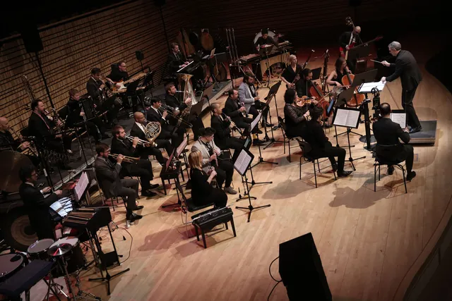 a male conductor in a black suit directs an orchestra on the concert hall stage. 