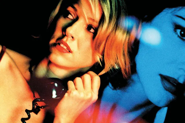 A white woman with blonder hair looking over her shoulder on the phone. An image of another woman is projected behind her in blue.