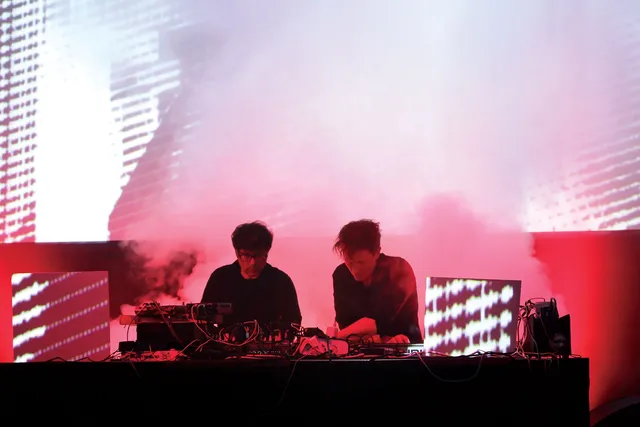 Two men DJing on a stage in front of was of red light and theatrical smoke. 