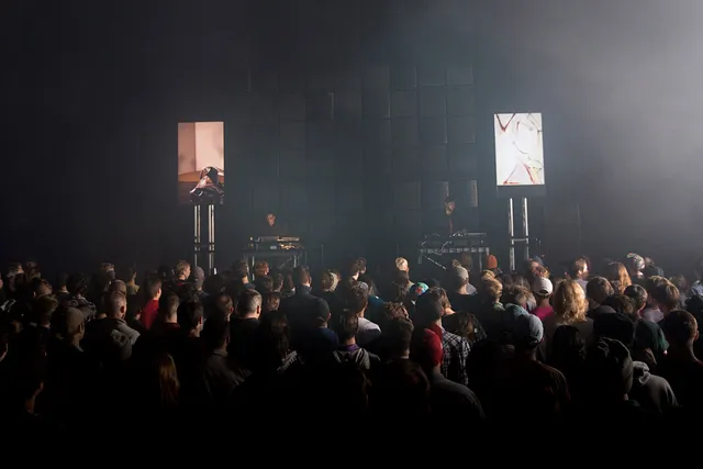 A crowded concert in a black box studio. People pack the stage watching oneohtrix. 