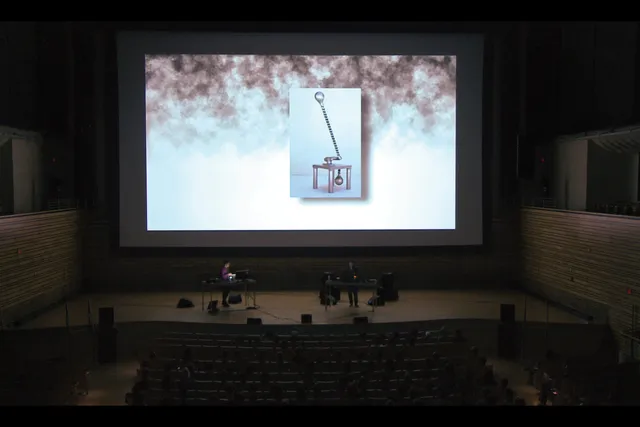 Two people behind desks and in discussion on stage infant of a large screen projecting the image of an abstract sculpture, resembling a low table with a long handle.