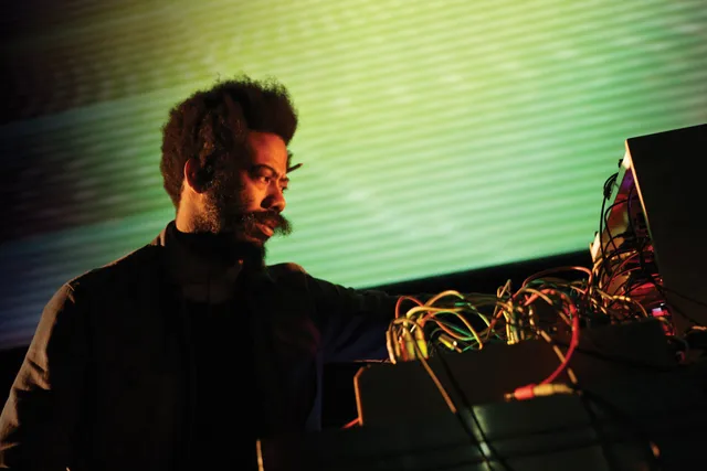 A Black man with a prominent mustache in front of a green and yellow projection examining various colored wires. 