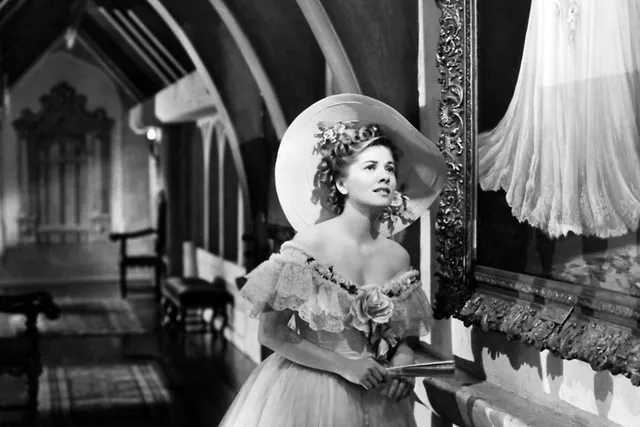 A southern woman dressed in civil war era clothing standing in an ornately decorated hallway, looking pensively at a painting.