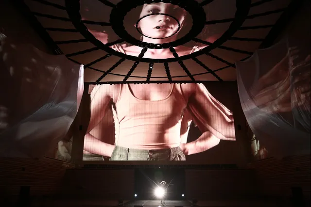 Projections of a female on the concert hall stage. Her head on the ceiling, torso on the back wall and arms outstretched on flowing fabric hung from the sides of the stage. 