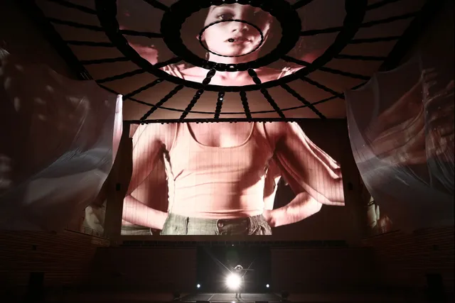 Projections of a female on the concert hall stage. Her head on the ceiling, torso on the back wall and arms outstretched on flowing fabric hung from the sides of the stage.
