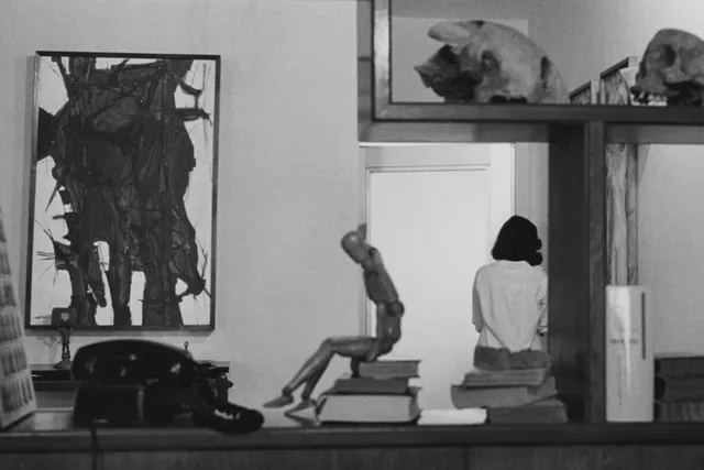 A table parallel to the view of the camera with books, a wooden artists dummy, and a rotary phone. A neatly decorated apartment with an AbEx painting can be seen in the background. A woman stand with back to the viewer leaning against the wall. 