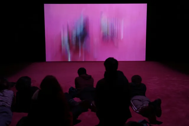A small silhouetted audience laying on a pink rug watching a projection of pink brush strokes in a dark room.