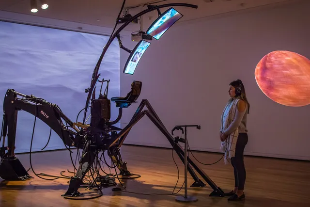 A woman wearing headphones looking at two screens held by a spider-like apparatus or sculpture in blank room with a projection of the ocean and a planet.
